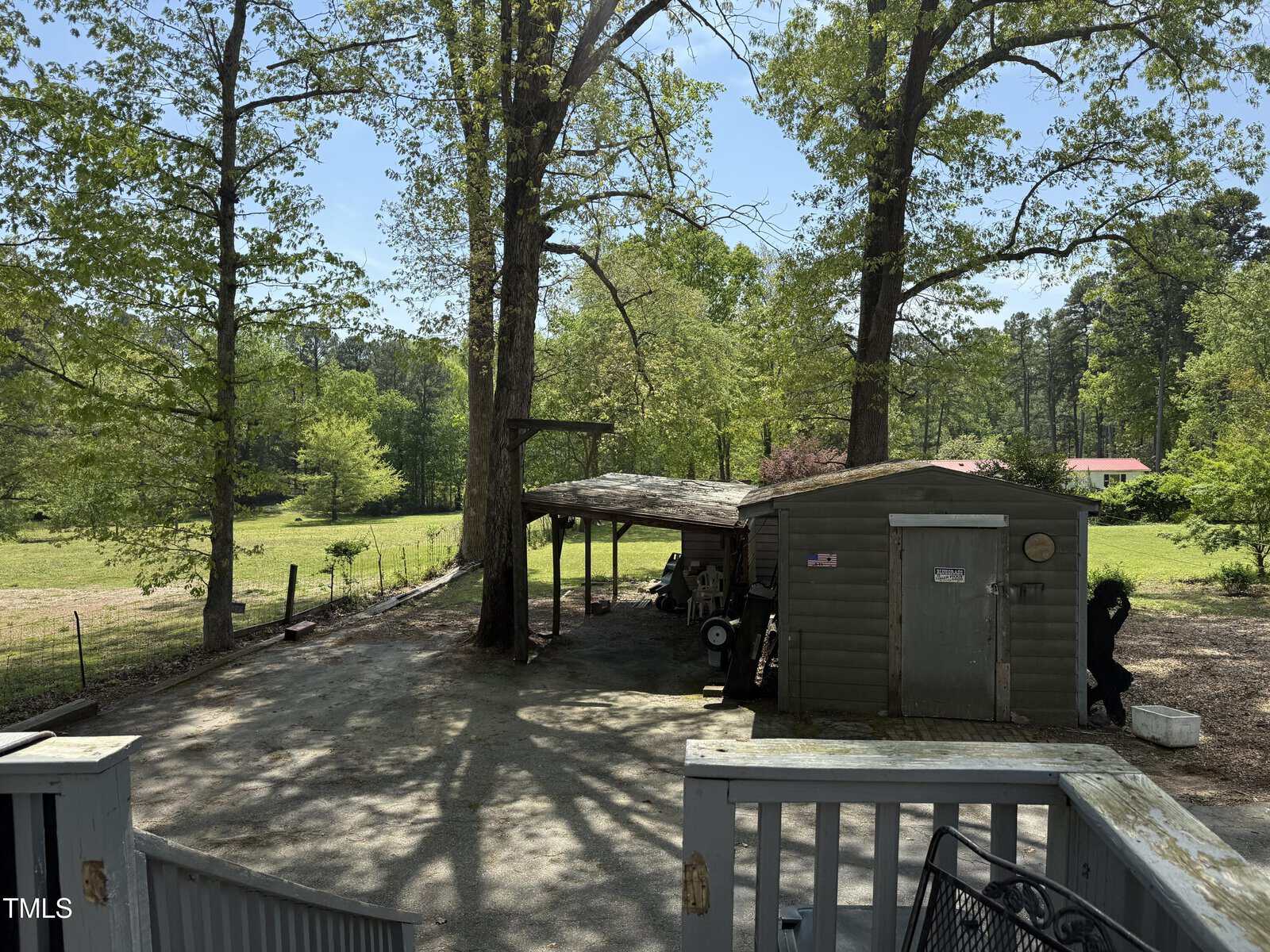 A small wooden shed with an attached covered area sits in a fenced backyard surrounded by trees and greenery on a sunny day. Gardening tools and equipment are visible under the covered area.