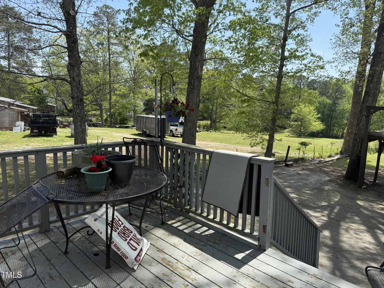 A wooden deck with a round metal table holding flower pots, a chair, a rolled rug, and hanging planters. The deck overlooks a grassy yard with trees, a shed, a truck, and a partially visible road.