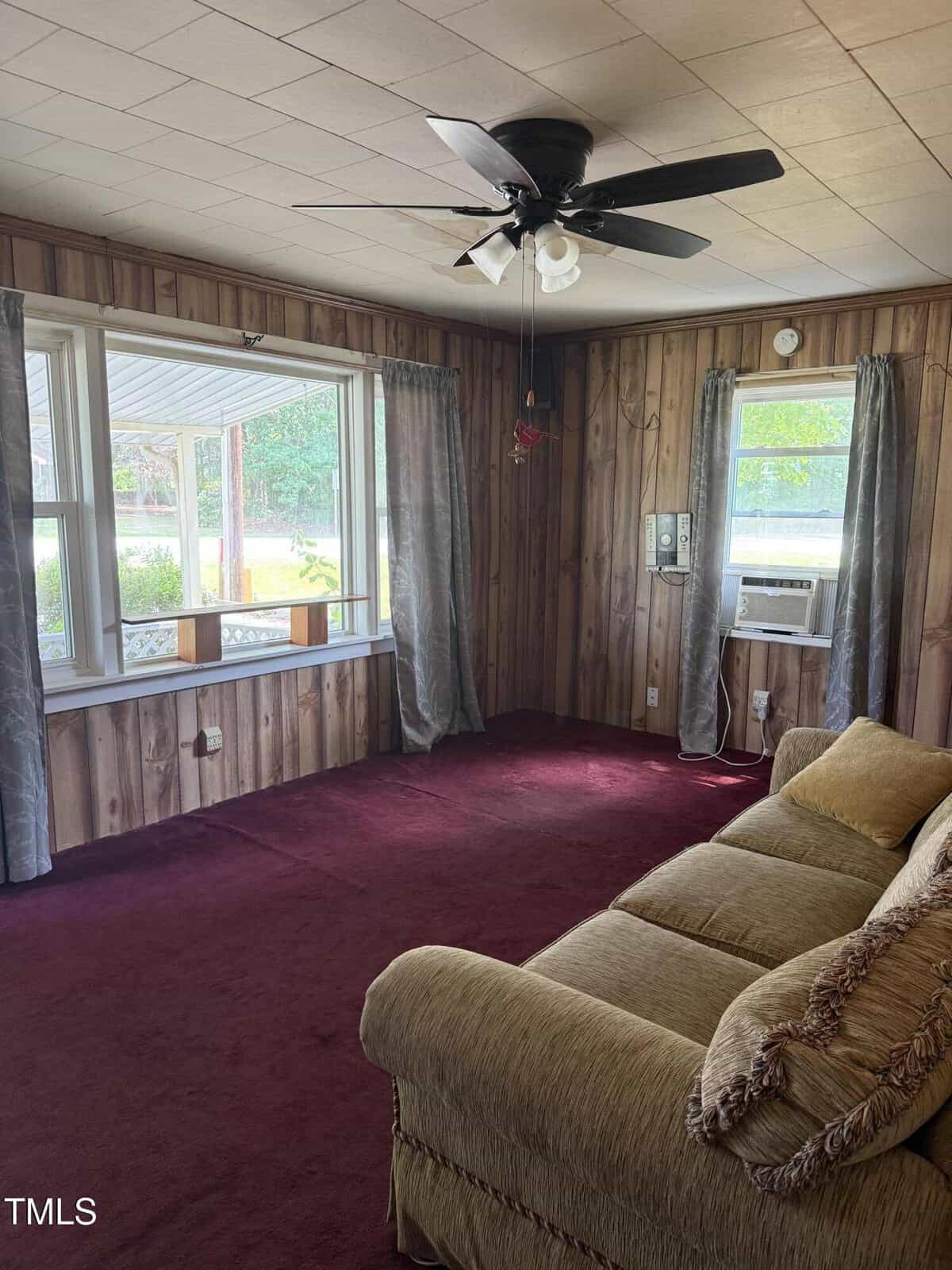 A living room with wood-paneled walls, maroon carpet, a tan couch, large windows with gray curtains, a ceiling fan with lights, and a window air conditioner. Greenery is visible outside the windows.