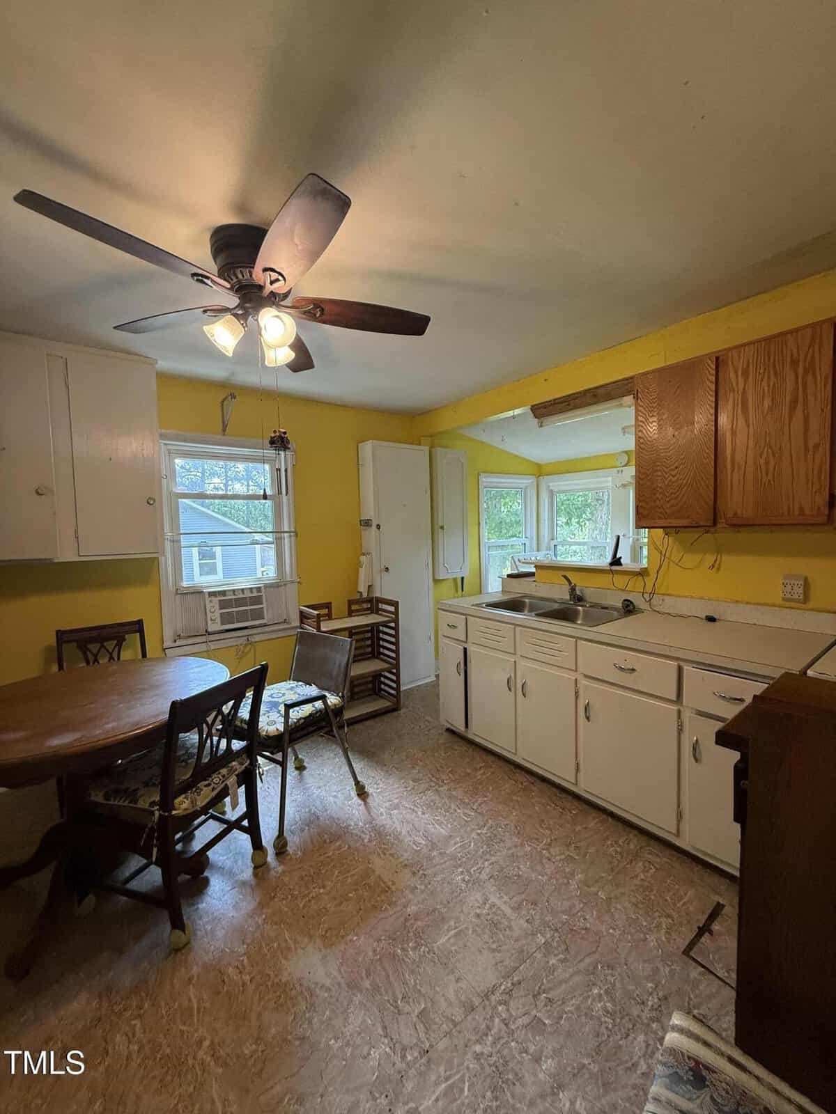 Older kitchen with yellow walls, white cabinets, brown ceiling fan with lights, round table with four chairs, and a window overlooking a sunroom. Countertops are cluttered, and flooring appears worn.