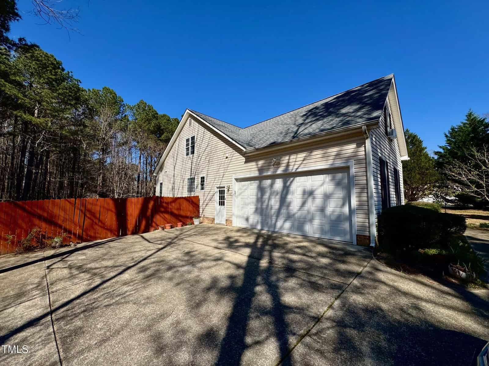 A beige two-story house with a double garage sits next to a concrete driveway. Shadows from leafless trees cross the driveway, and a tall red wooden fence runs along the side. Pines and shrubs are in the background.