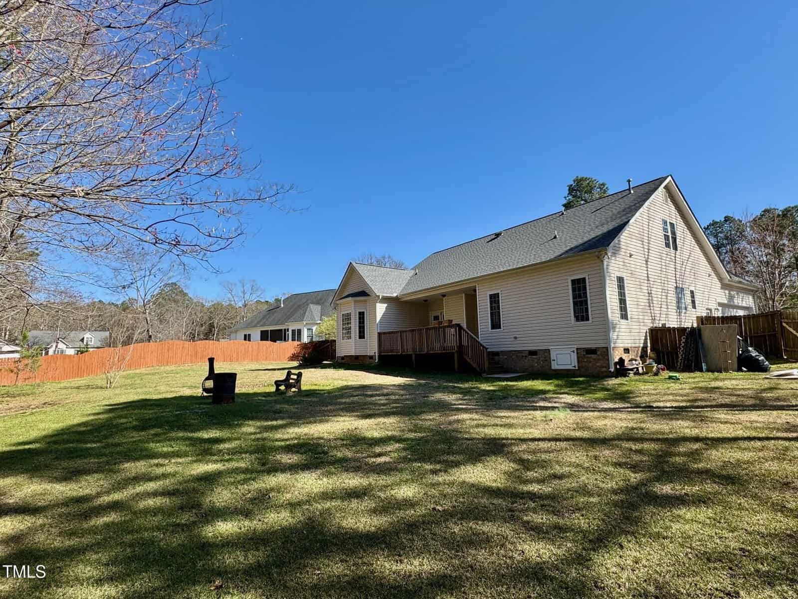 A spacious fenced backyard with a grassy lawn, two outdoor chairs around a firepit, and a beige house with a covered back porch under a clear blue sky.