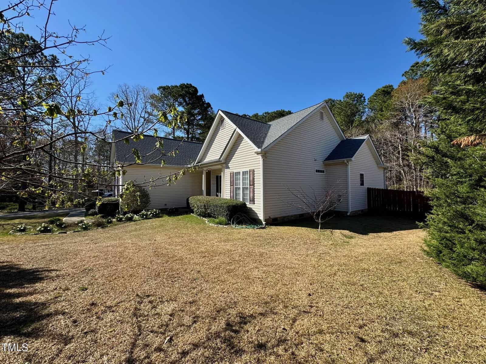 Single-story beige house with dark gray roof, white trim, and attached garage. The front yard has dry grass, shrubs, and trees, with a driveway on the left. The sky is clear and blue.