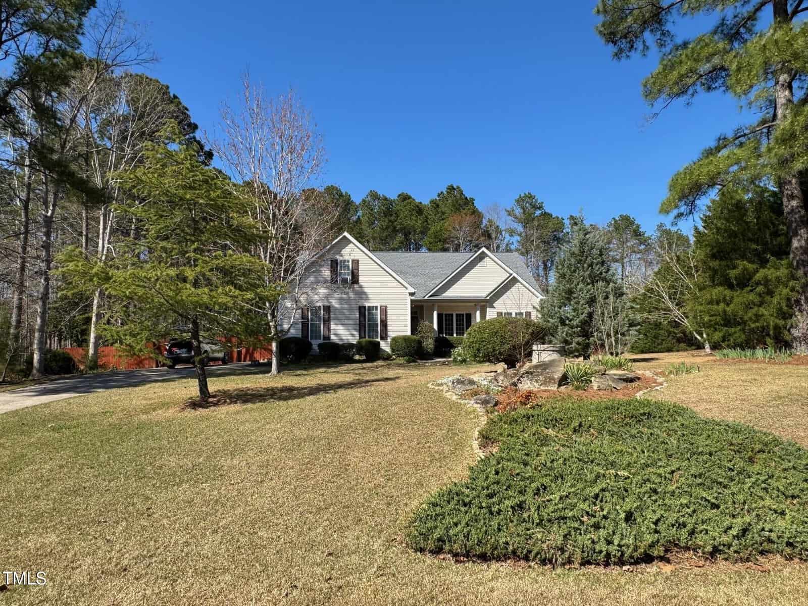 A beige single-story house with a grey roof sits amid a spacious, neatly landscaped yard with green shrubs, trees, and a clear blue sky in the background.