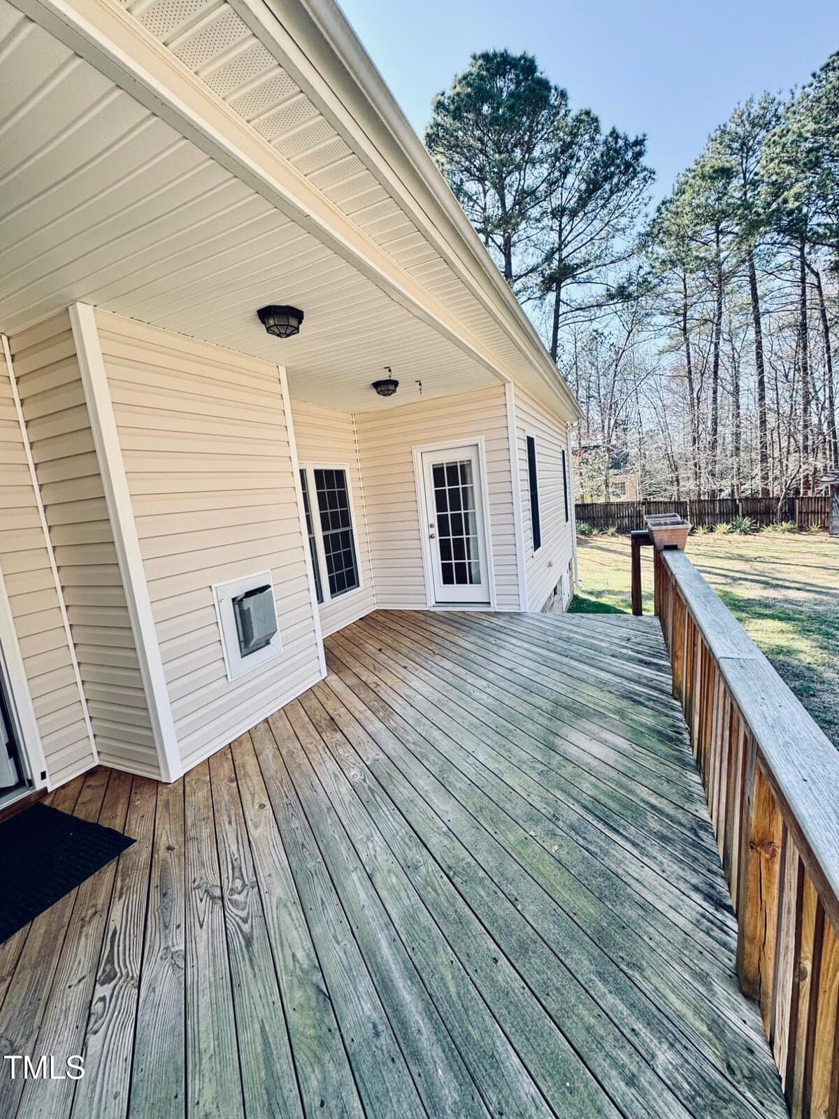 A wooden deck with a railing attached to a light-colored house, featuring two exterior doors, wall-mounted lights, and surrounded by a grassy yard with tall trees in the background on a sunny day.