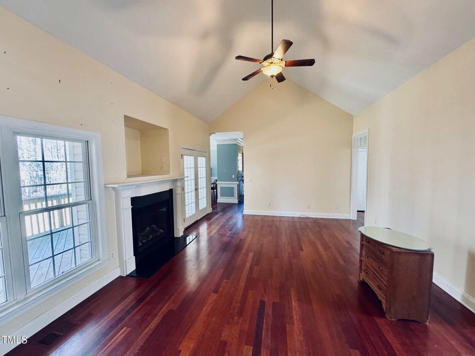 A spacious living room with hardwood floors, cream-colored walls, a ceiling fan with light, fireplace, large windows, and a small wooden dresser. The room is bright and opens to another area through a wide doorway.