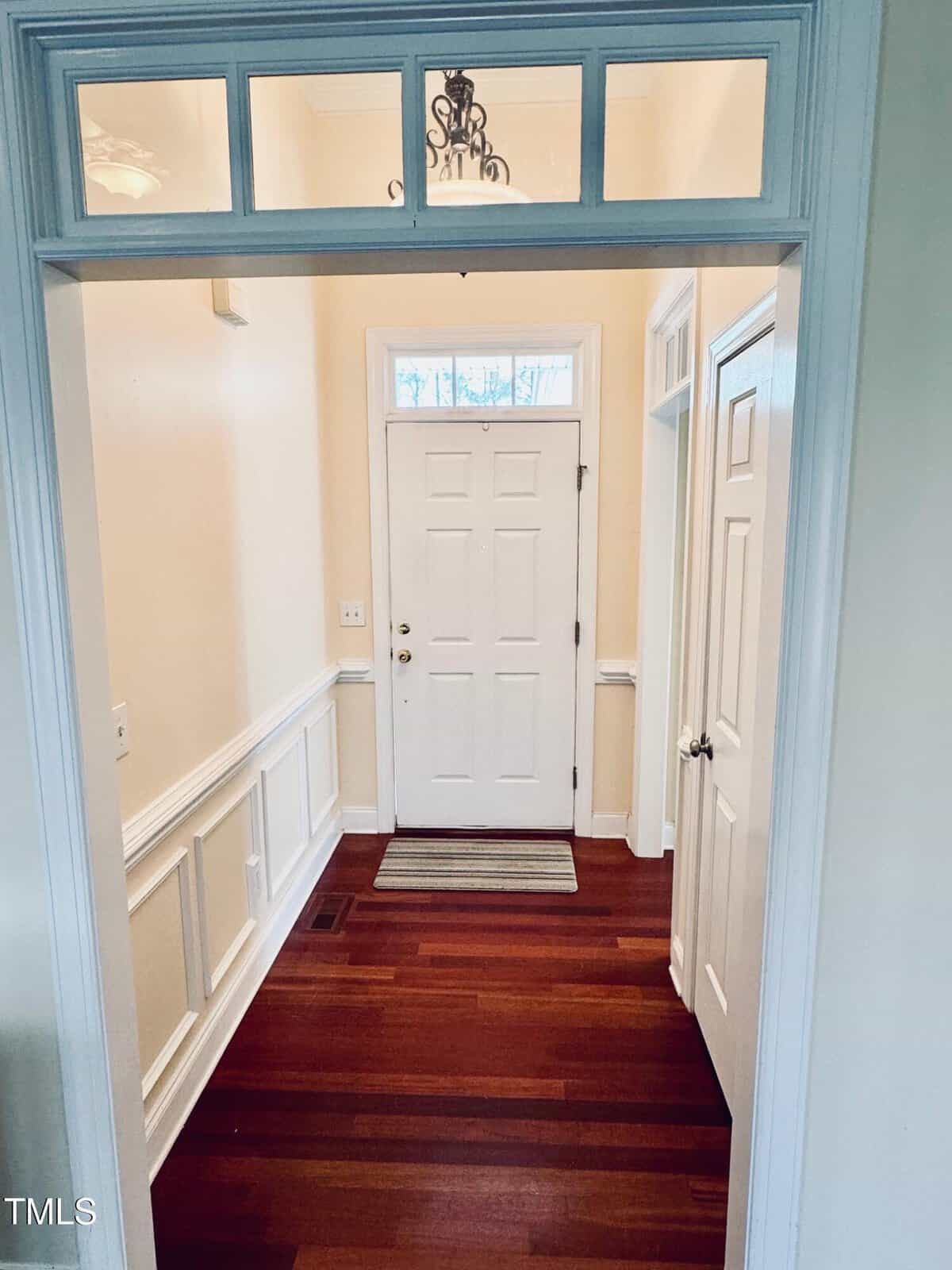 A bright entryway with a white front door, small upper window, beige walls, white trim and panel wainscoting, rich wood floors, a striped rug, and a small chandelier overhead.
