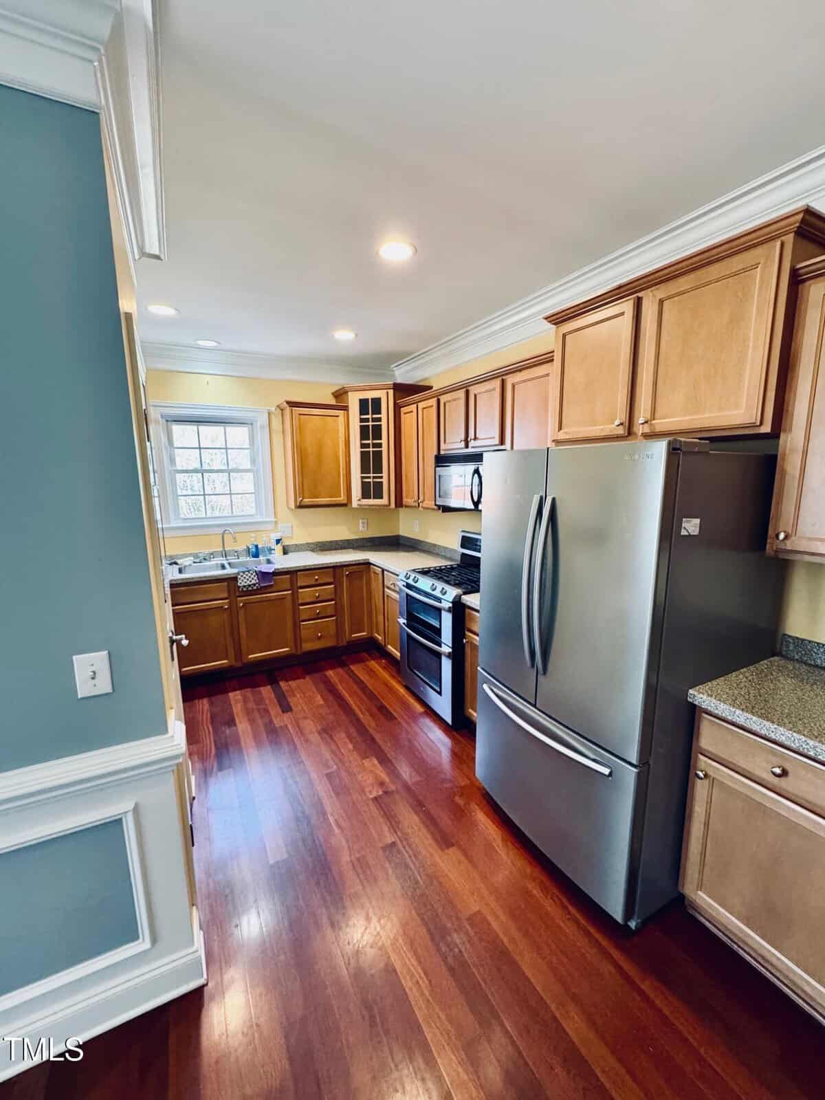 Modern kitchen with stainless steel appliances, light wood cabinets, granite countertops, and hardwood floors. A window above the sink provides natural light. Recessed ceiling lights illuminate the space.