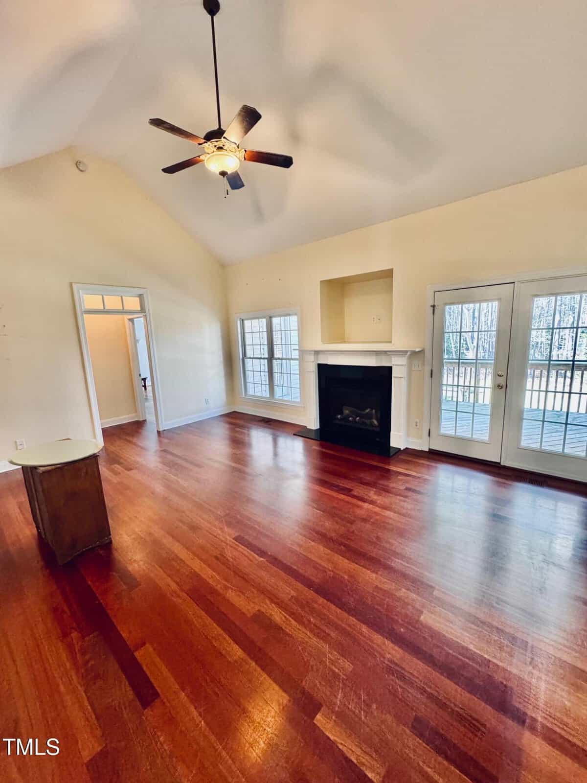 Spacious living room with high ceiling, ceiling fan, hardwood floors, fireplace, and large windows and doors opening to a deck with wooded views.