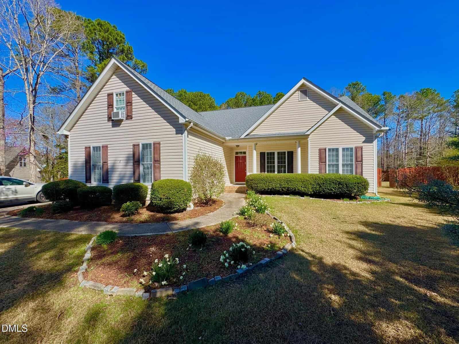 Single-story beige house with red shutters, a central red door, gray roof, and a front yard with trimmed bushes, small flower beds, and a concrete walkway. Nestled at 20 Willows Den Court in Youngsville, the home is surrounded by trees under a clear blue sky.