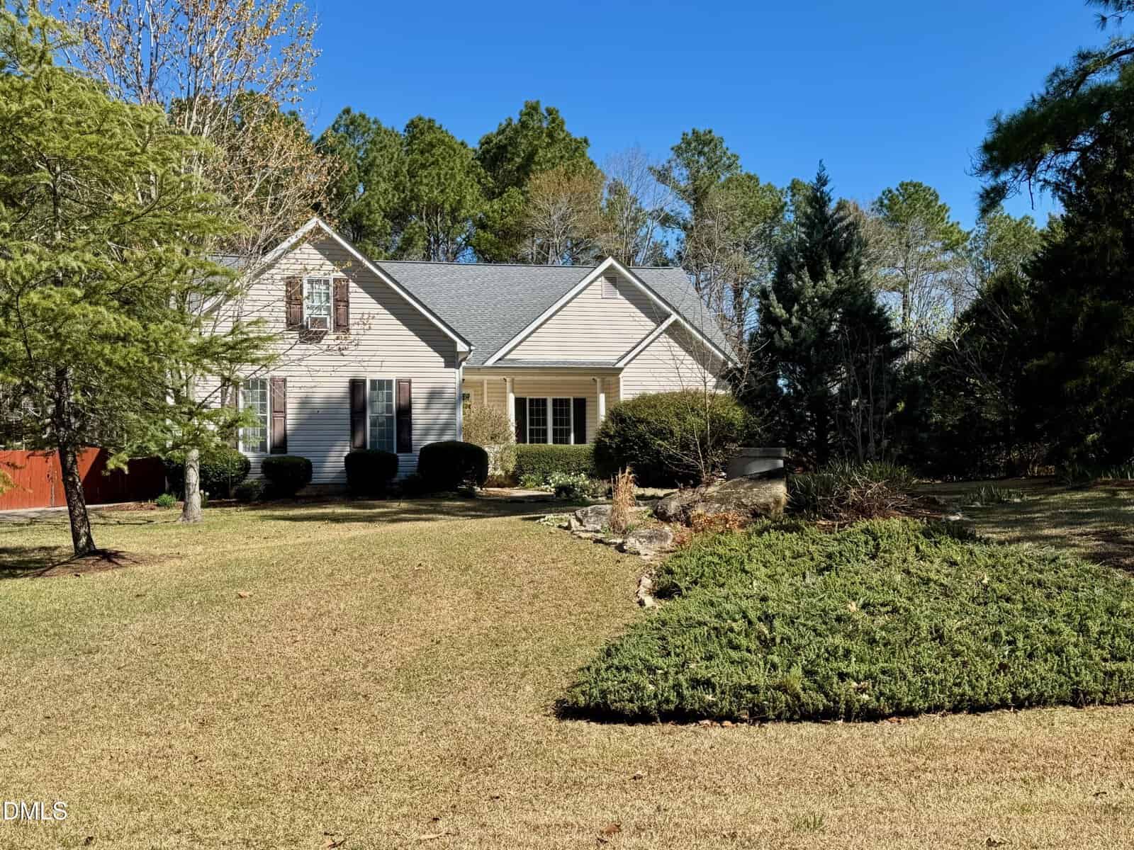 Single-story beige house with gray roof, white trim, and dark shutters, surrounded by neatly trimmed bushes, trees, and a large, well-maintained lawn on a sunny day.