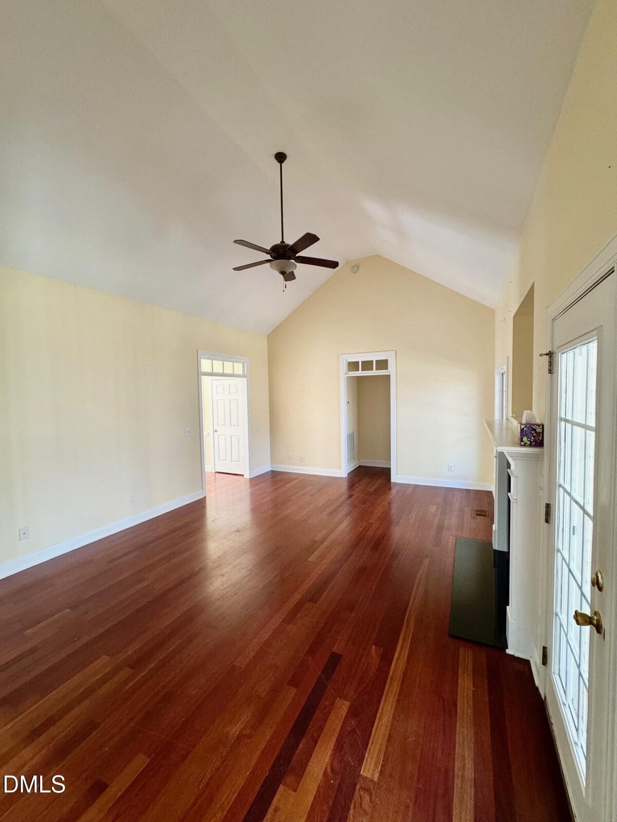 A spacious, empty living room with vaulted ceiling, ceiling fan, hardwood floors, pale yellow walls, a fireplace, and glass doors leading outside. Natural light fills the space from multiple windows.