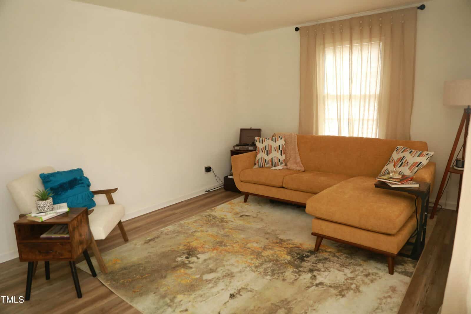 A living room with a mustard-colored sectional sofa and patterned pillows, a beige armchair, and a wooden side table with books. A large window with sheer curtains is in the background, and a patterned rug covers the floor.
