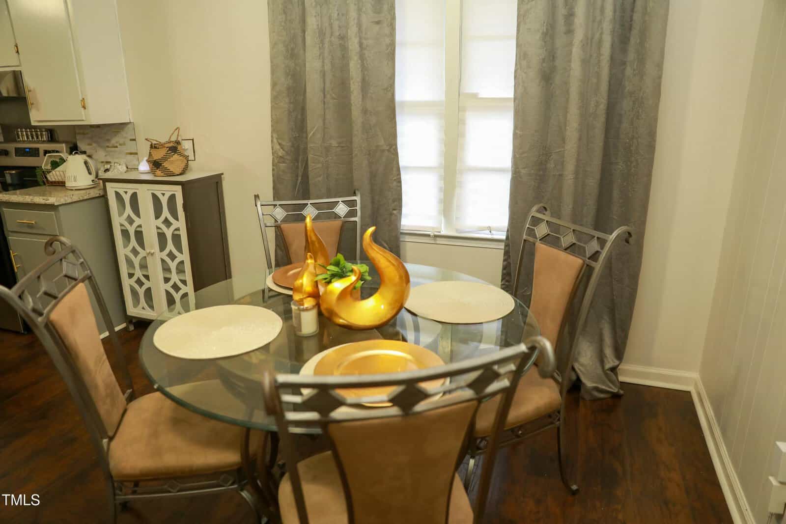 A dining area features a round glass table with four metal chairs that have tan cushions. The table is set with beige placemats and gold plates, and a decorative arrangement sits in the center. Gray curtains cover the window in the background.