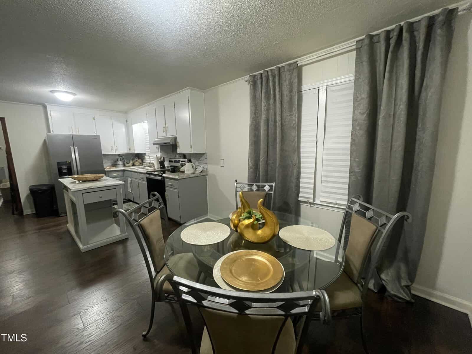 A kitchen and dining area with dark wood flooring. There's a round glass dining table with four chairs, set with placemats and a decorative centerpiece. The kitchen features white cabinets, stainless steel appliances, and a small island.
