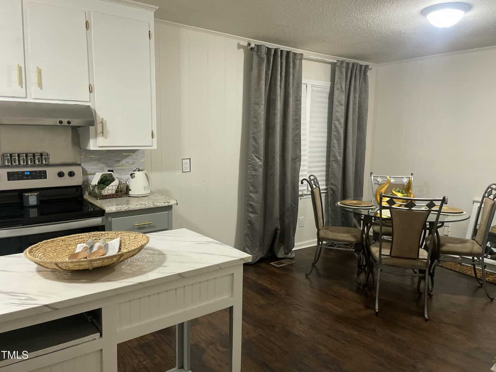 A kitchen with white cabinets, a stainless steel oven, and a countertop with a wicker tray. A small dining area with a round table and four chairs is visible. Gray curtains cover the window. The flooring is dark wood. A ceiling light is on.