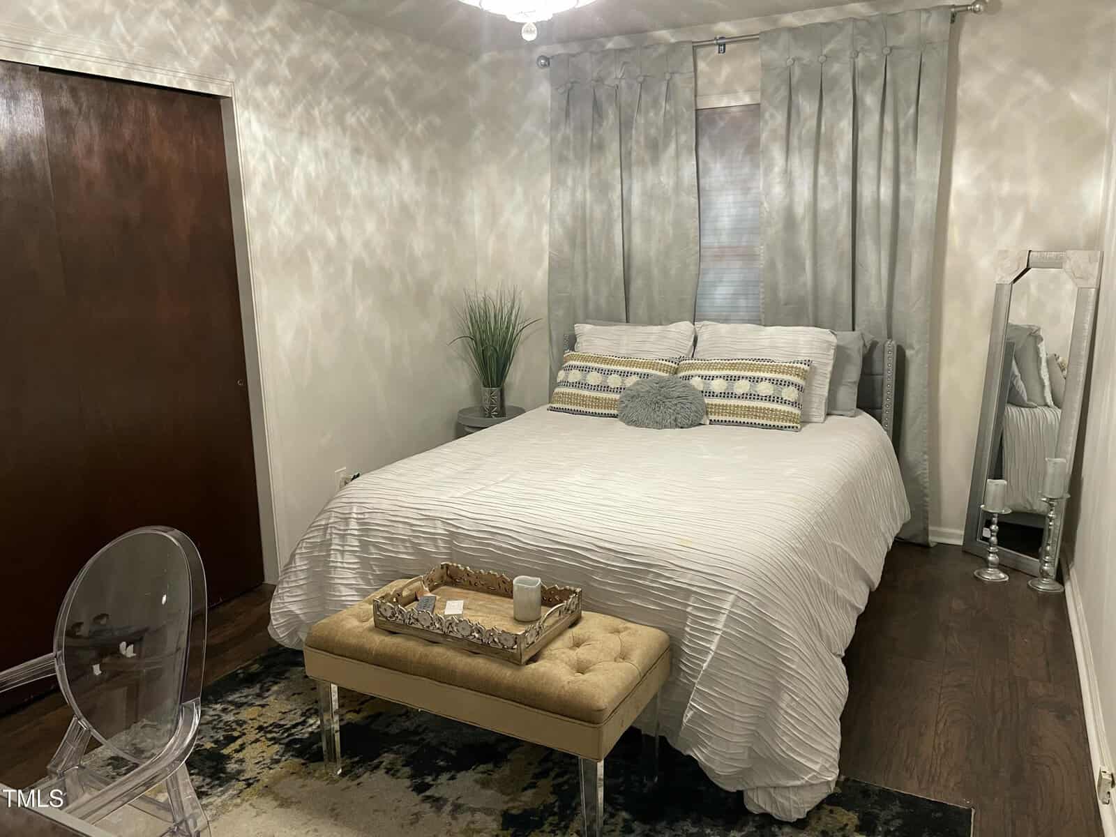 A bedroom with a double bed featuring a white comforter and decorative pillows. A wooden bench at the foot holds a tray with candles. A large mirror leans against the wall, and a small plant sits on a nightstand. A transparent chair is in the foreground.