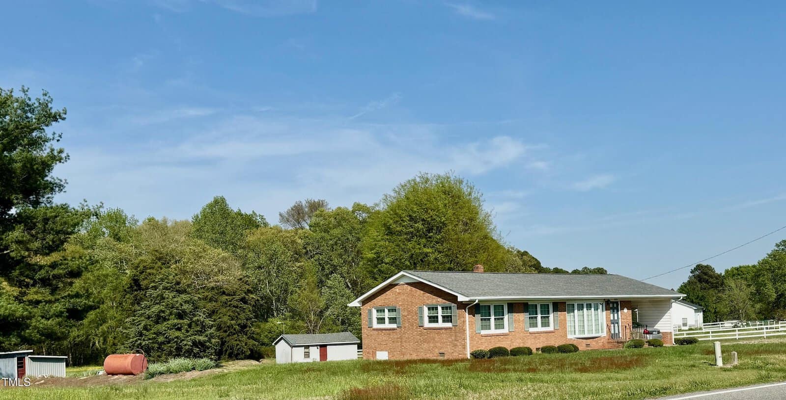 A red-brick single-story house with a gray roof is surrounded by greenery and trees. A white fence runs along the side, with a red barrel and outbuildings visible in the yard. The sky is clear and blue.