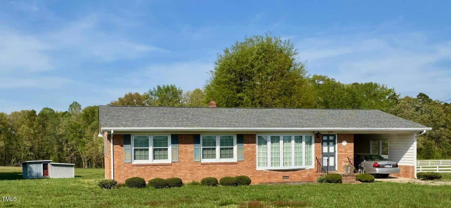 A single-story brick house at 1829 Stewart Farm Road features a shingled roof and large windows. A small covered carport on the right houses a parked car, while the grassy yard is dotted with shrubs. In Henderson, a quaint shed sits against a backdrop of trees.