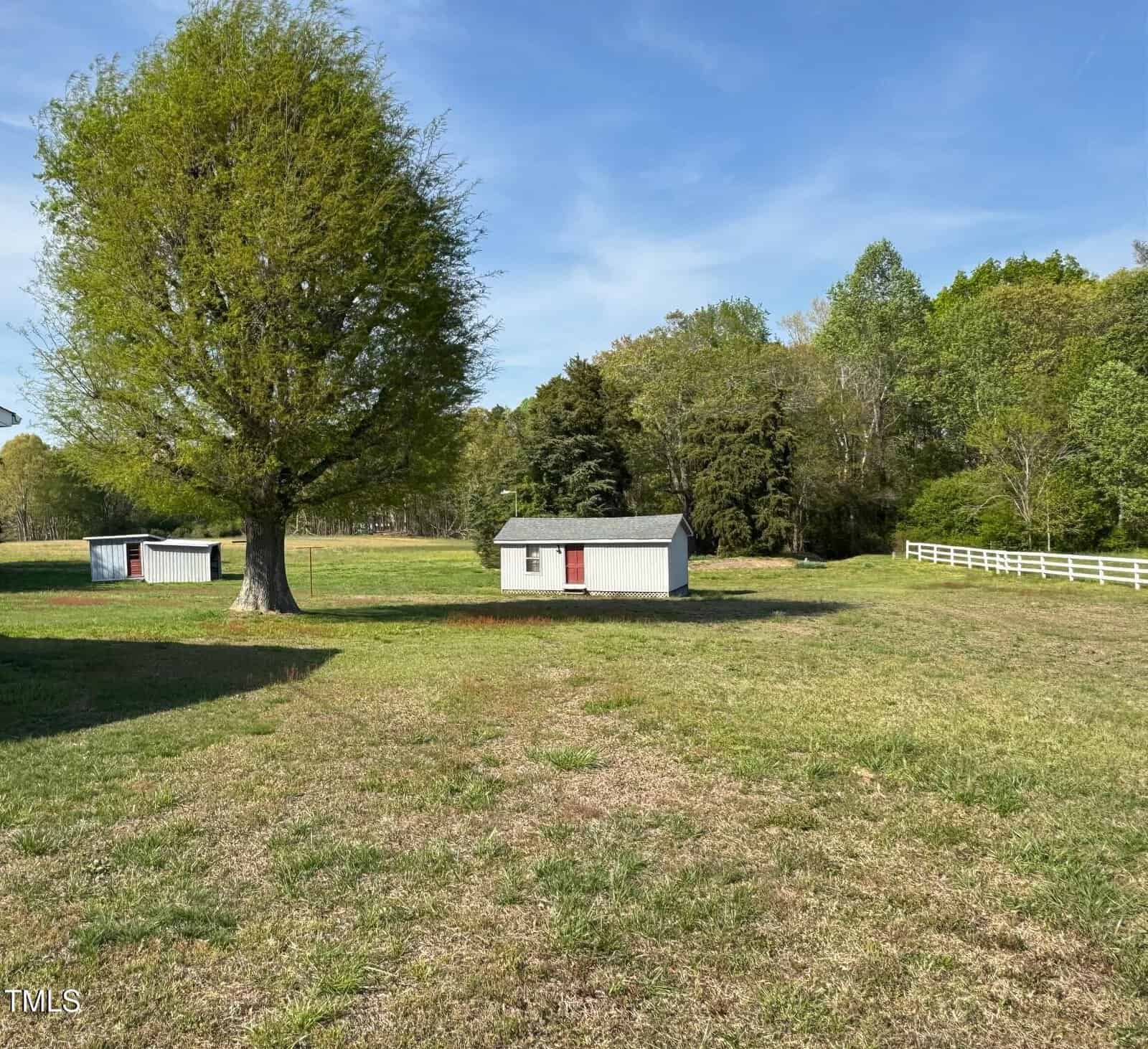 A grassy field with two small white sheds under a large tree. The sheds have red doors and green roofs. A white fence runs along the right side, and trees with green foliage are in the background under a blue sky.