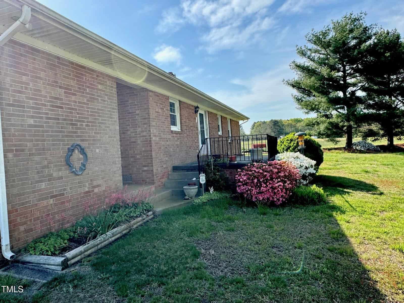 A red brick house with a small porch and steps, surrounded by a lawn. A neatly trimmed garden bed with pink and white flowering bushes is visible in the foreground. A large evergreen tree is to the right, and the sky is partly cloudy.