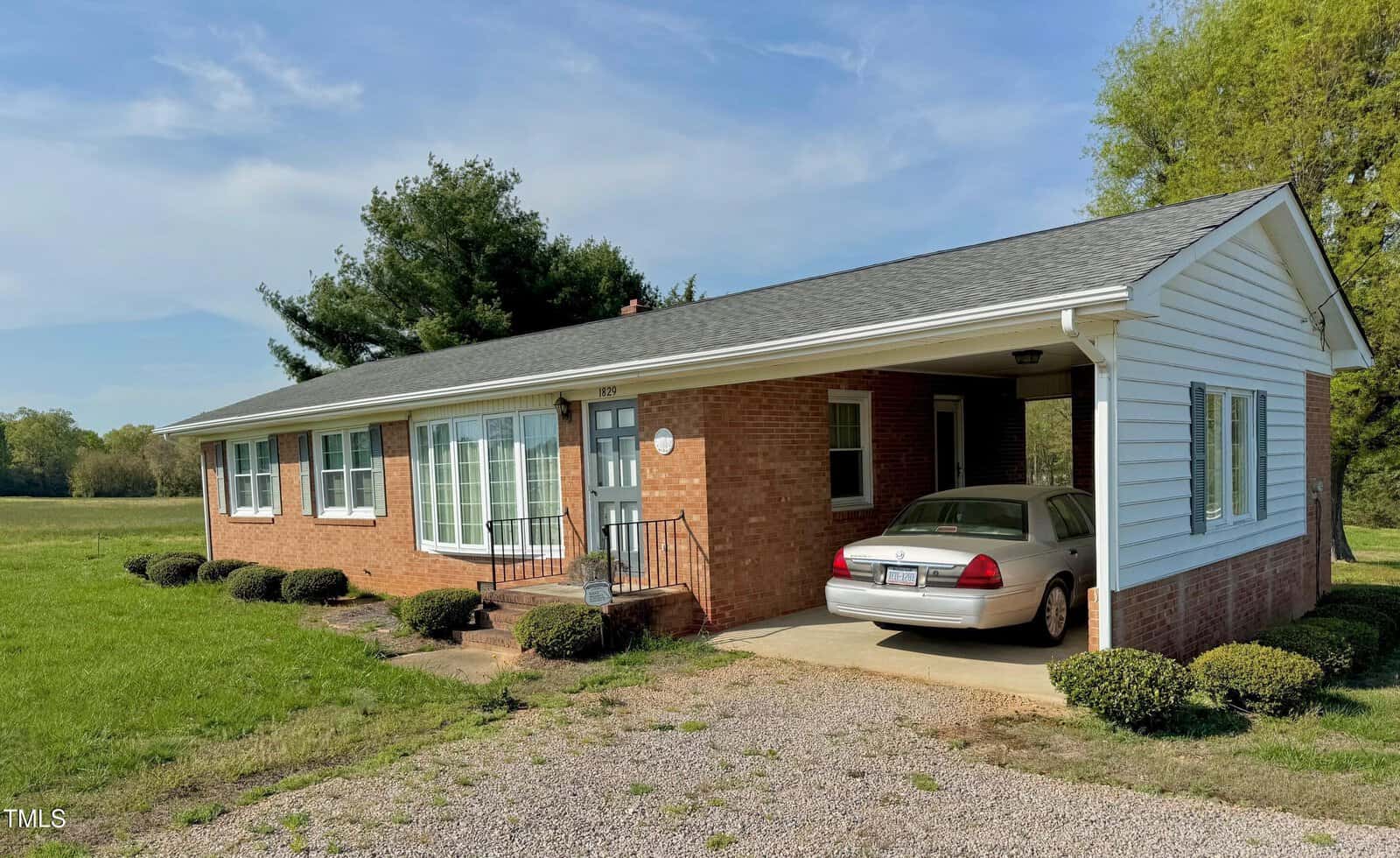 Single-story brick house with a gray shingled roof and white trim. A car is parked under an attached carport. The house has several windows and small bushes lining the front. The sky is clear, and a grassy field surrounds the building.