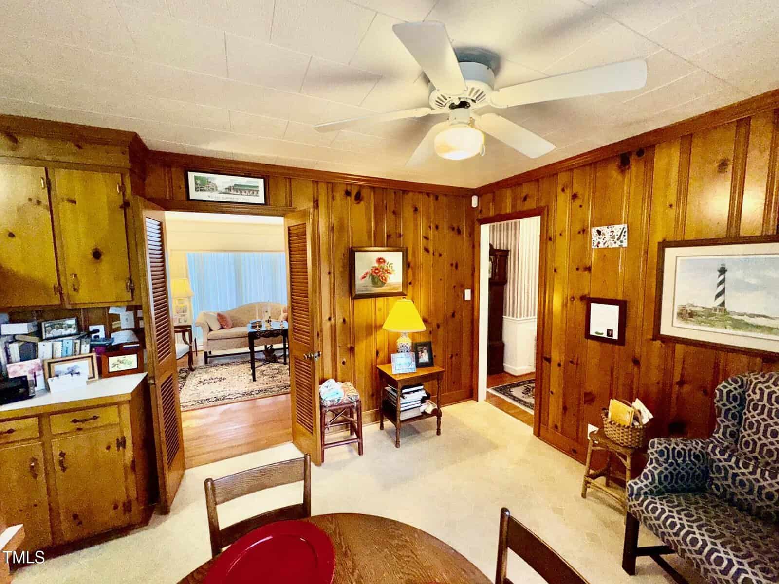 A wood-paneled room with a ceiling fan, built-in cabinets, and a table with chairs. An open doorway leads to a living area, and artwork and books decorate the space.