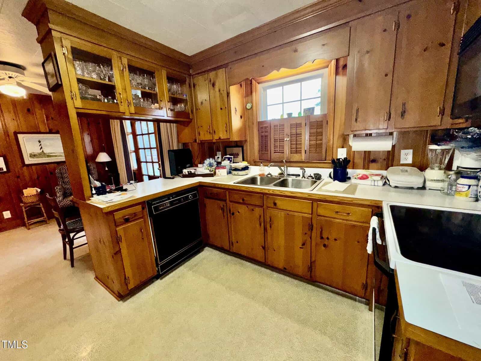 A kitchen with wood-paneled cabinets, a double sink under a window with wooden shutters, a dishwasher, a stove, countertop appliances, and an open shelving area with glasses above the counter.