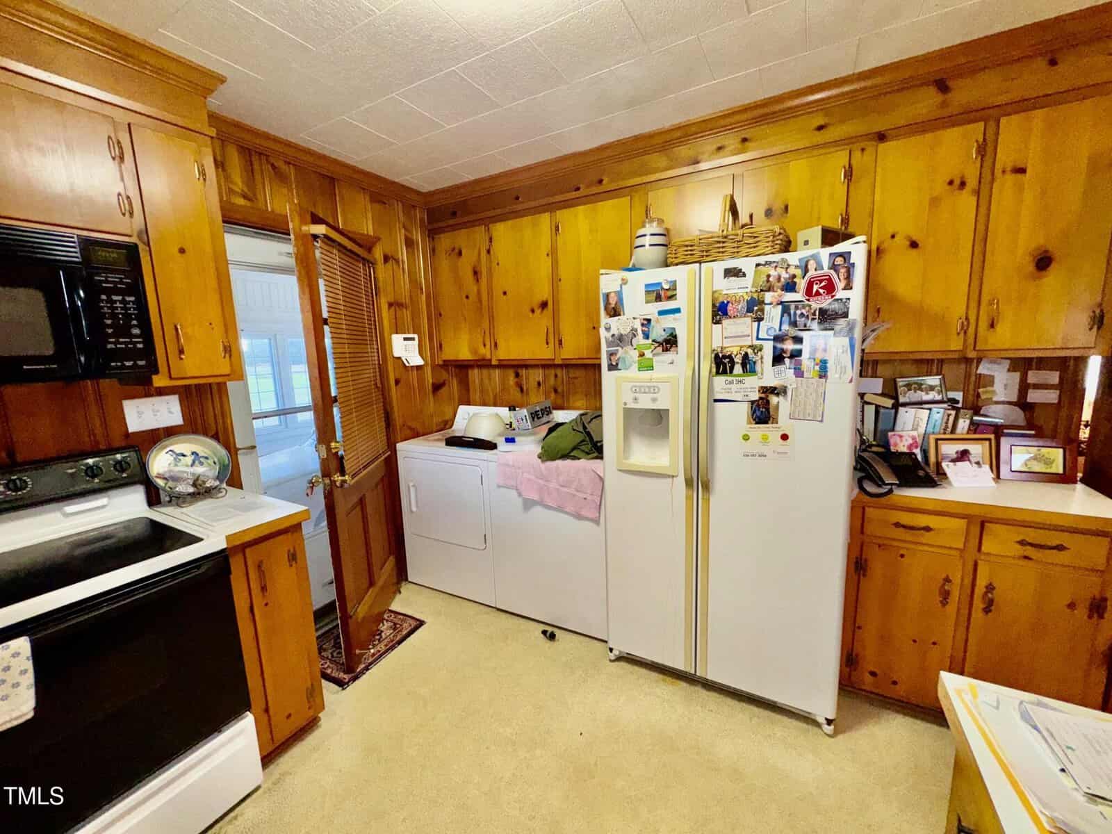 A kitchen with wood-paneled cabinets, a white refrigerator covered in photos and magnets, a countertop with various items, a white oven, a microwave, and a washer and dryer next to a wooden door.