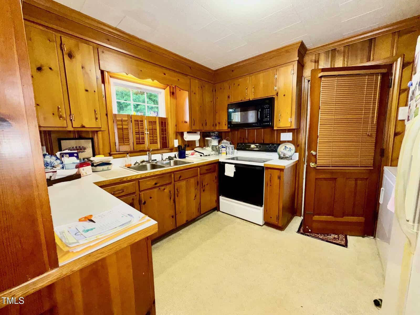 A kitchen with wood-paneled cabinets and walls, a white stove, a microwave, a double sink under a window with shutters, and a wooden door with blinds. Countertops have various kitchen items and papers.