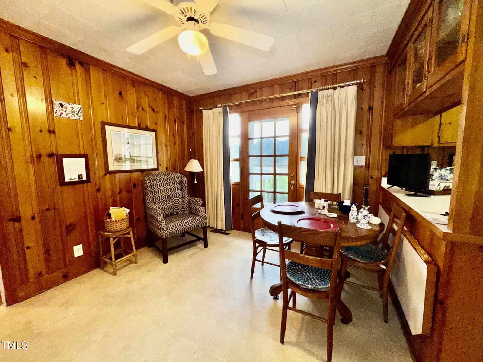 A wood-paneled room features a round dining table with four chairs, a patterned armchair, a floor lamp, wall art, a ceiling fan, curtained windows, and a door leading outside. A small television is on a side counter.
