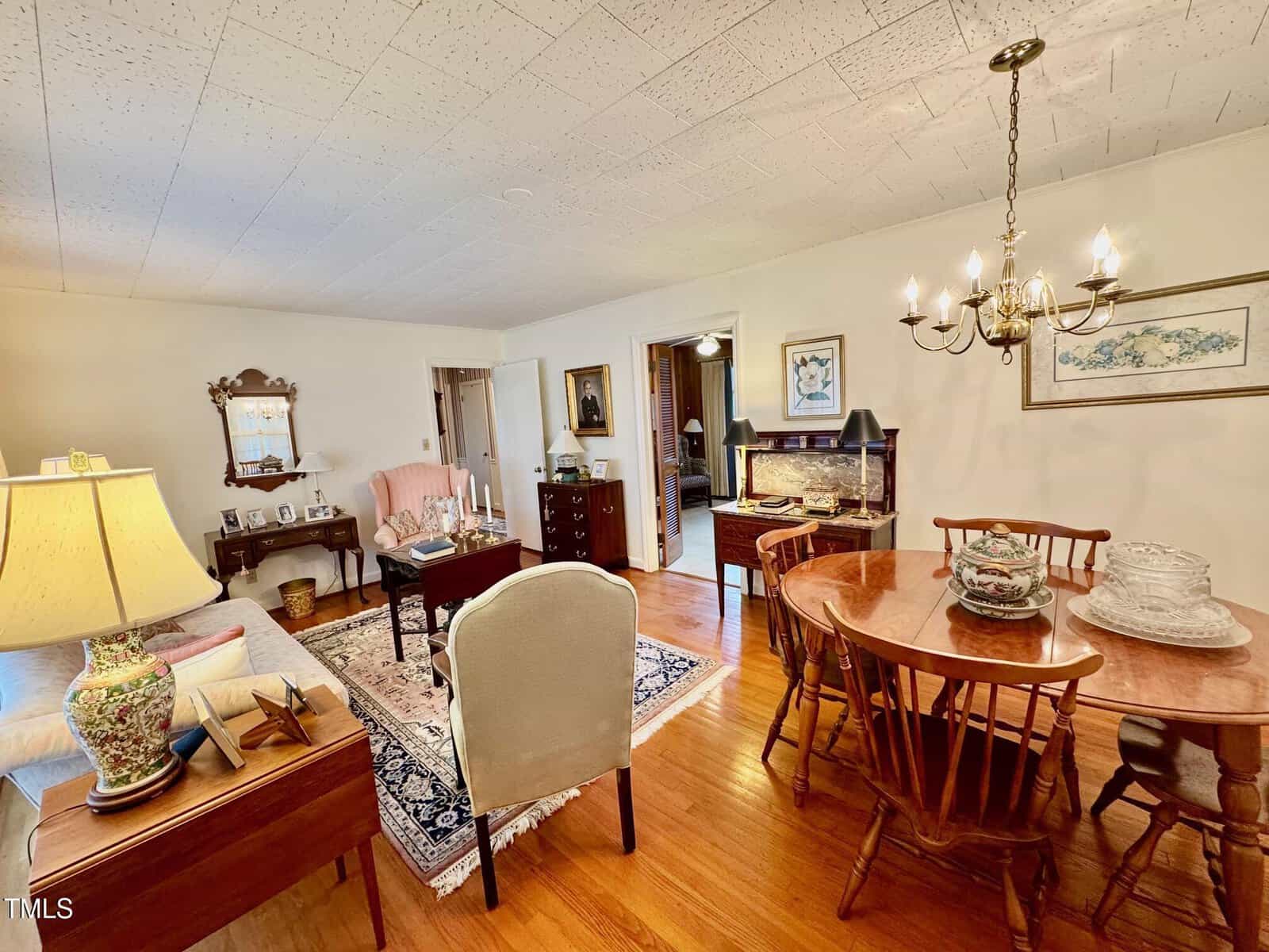 A traditional living and dining room features hardwood floors, a round wooden dining table with six chairs, a chandelier, armchairs, a sofa, framed artwork, and various decorative items on tables and shelves.