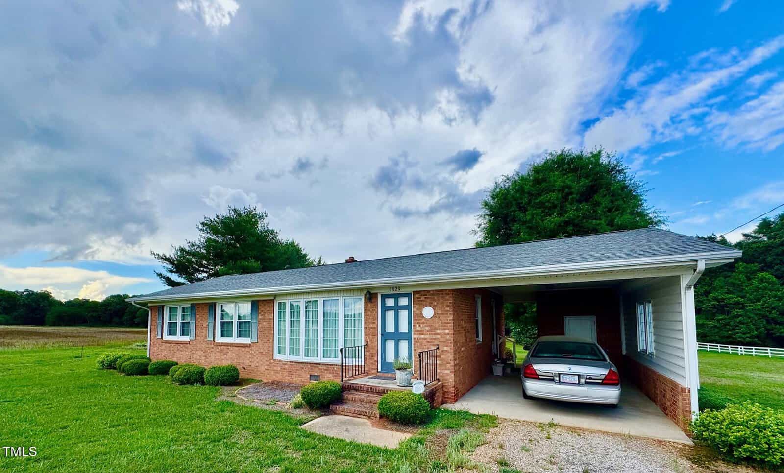 A one-story brick house with white trim and blue front door, featuring large windows and an attached carport with a white car parked under it. The house is surrounded by grass, trees, and an open sky with clouds.