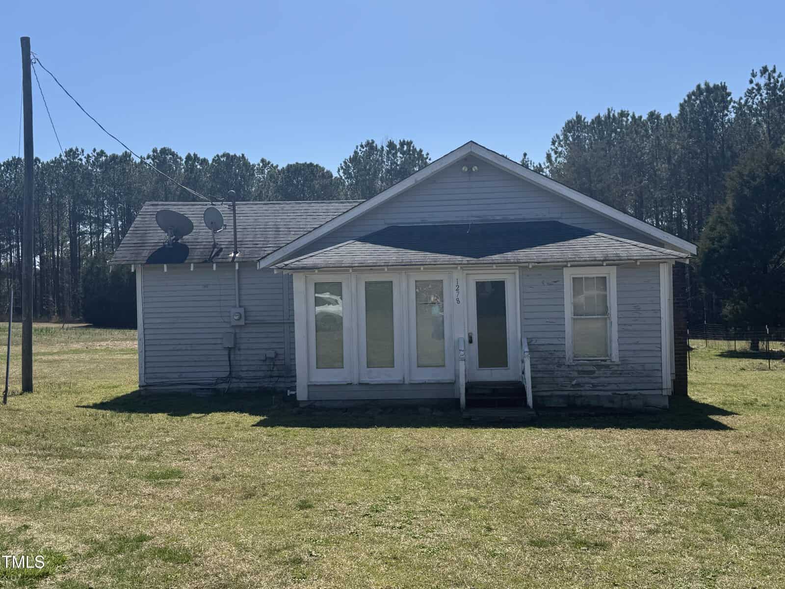A single-story, weathered gray house with a small front porch, large front windows, and a shingled roof, sits on a grassy lot with trees and a clear blue sky in the background. Two satellite dishes are attached to the side.