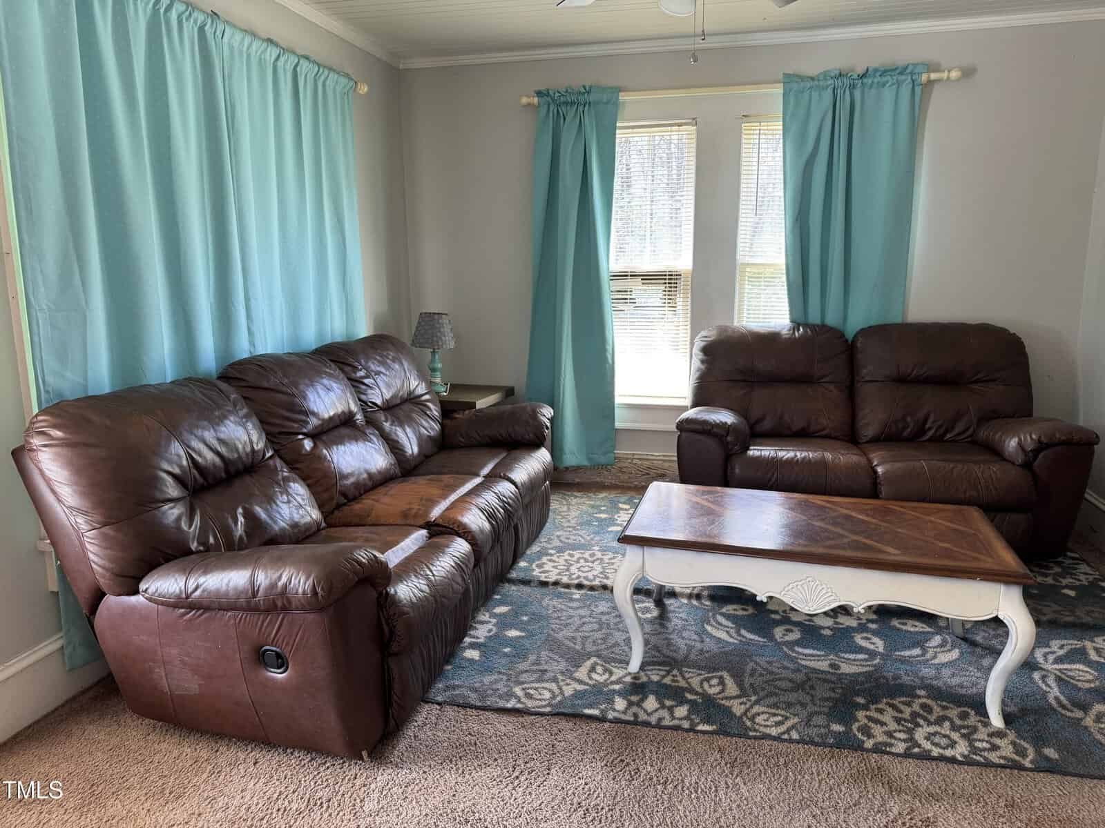 A living room with two brown leather sofas, a patterned rug, a wooden coffee table with white legs, light blue curtains, and a small table with a lamp in front of two windows.
