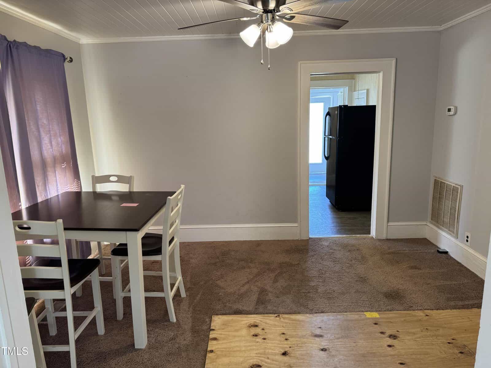 A small dining area with a dark table and four white chairs sits on brown carpet next to a window with purple curtains. A ceiling fan is above, and a kitchen with a black refrigerator is visible through an open doorway.