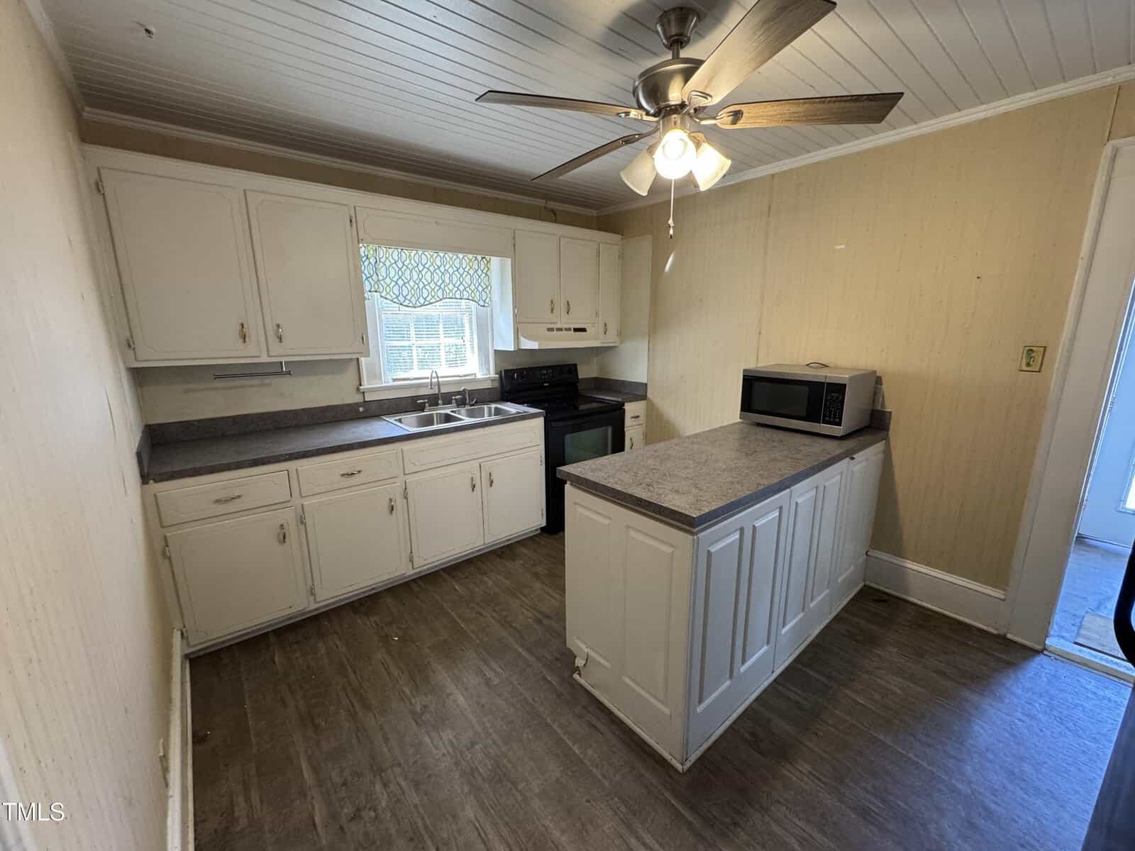 A kitchen with white cabinets, a gray countertop, a double sink under a window with a patterned valance, a black stove, a microwave on an island counter, wood-look flooring, and a ceiling fan with lights.