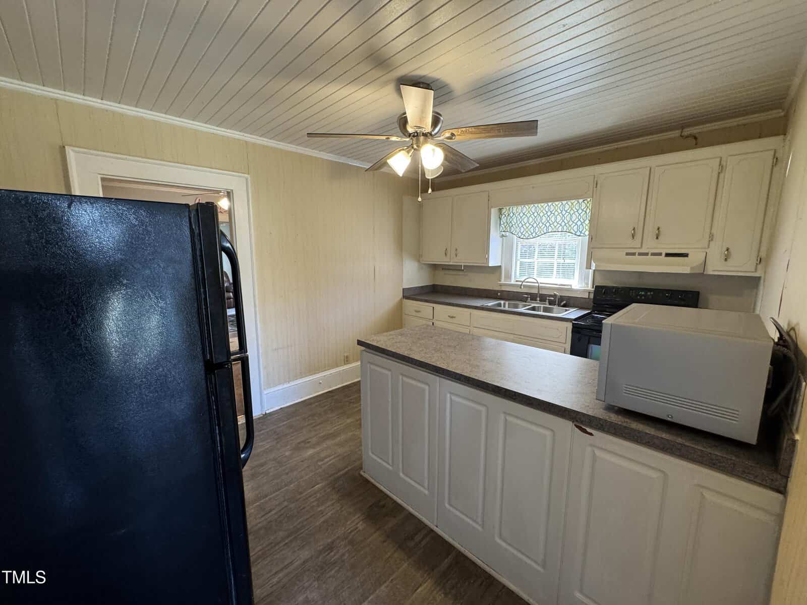 A kitchen with white cabinets, dark countertops, a black refrigerator, stove, microwave, and a ceiling fan with lights. There is a window above the sink and wood-patterned flooring.