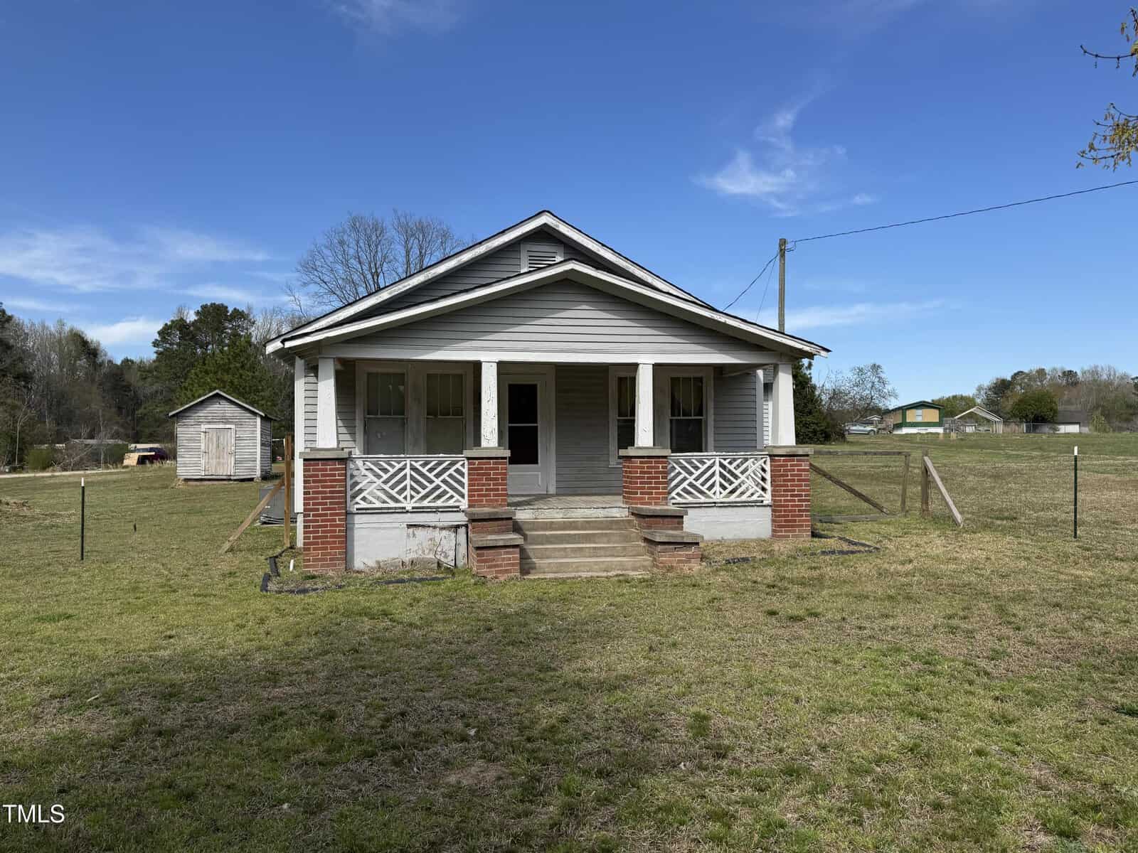 A small, single-story house with a porch, brick steps, and white railing stands in a grassy yard. There is a shed to the left and fields and trees in the background under a blue sky.