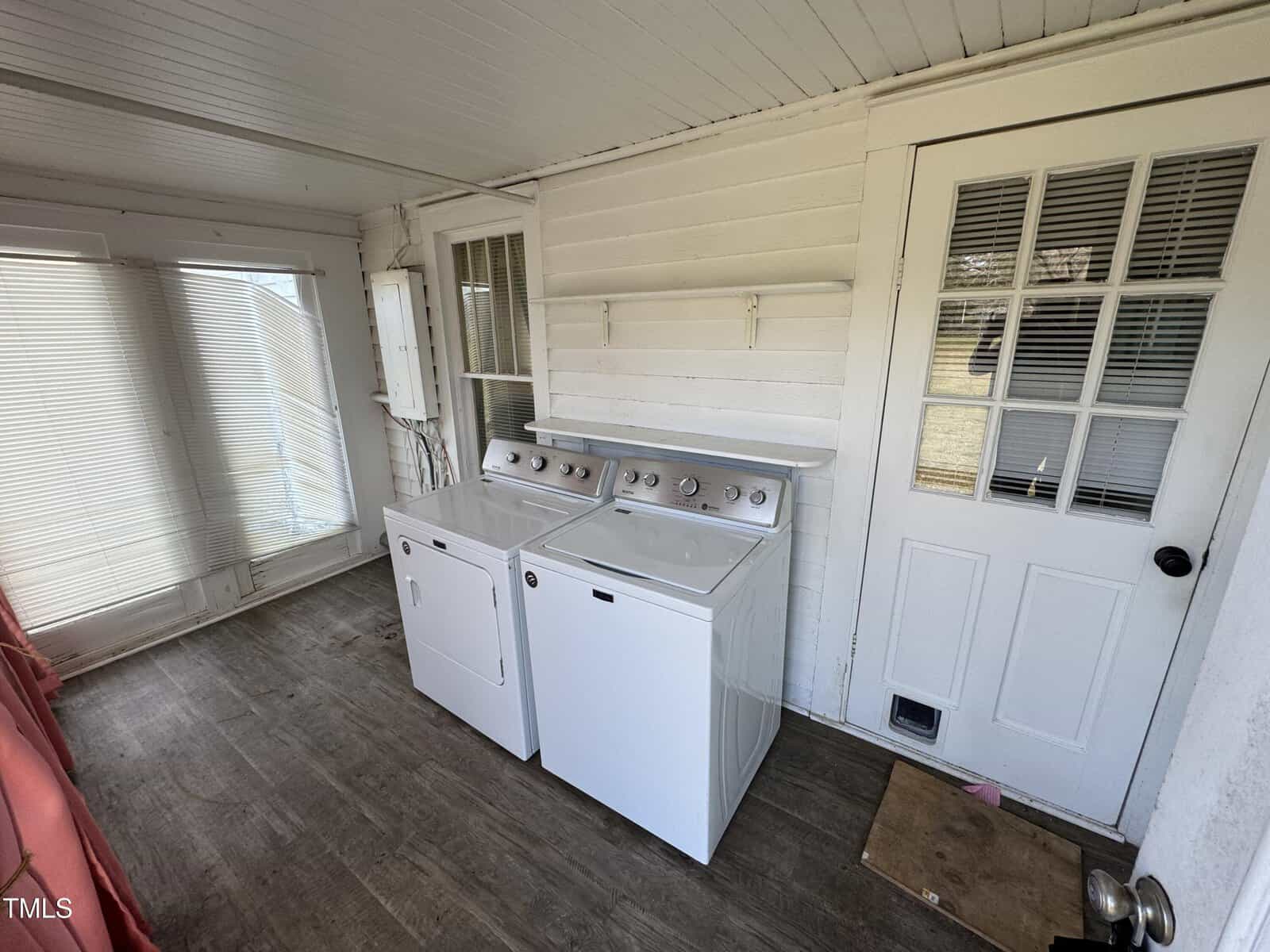 A laundry area with a white washer and dryer, white paneled walls, shelves above, a door with a window and pet door, large windows with blinds, and wood-look flooring.