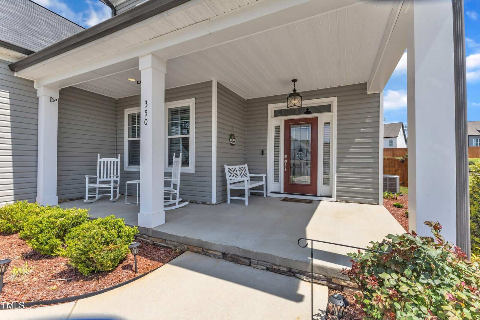 Gray house with white trim features a covered front porch, two white rocking chairs, a white bench, light fixture, and glass front door. Small shrubs and mulch line the walkway leading to the entrance.