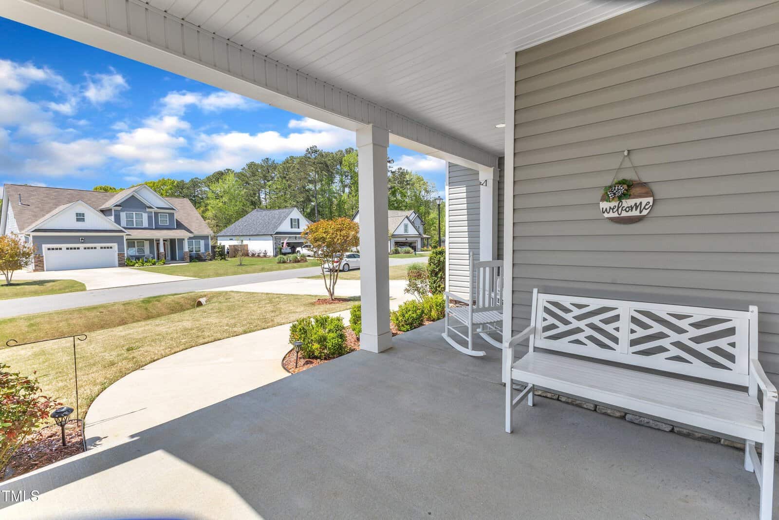 A covered front porch with a white bench, a “Welcome” sign on the wall, and a view of a suburban neighborhood with houses, lawns, trees, and a blue sky with scattered clouds.