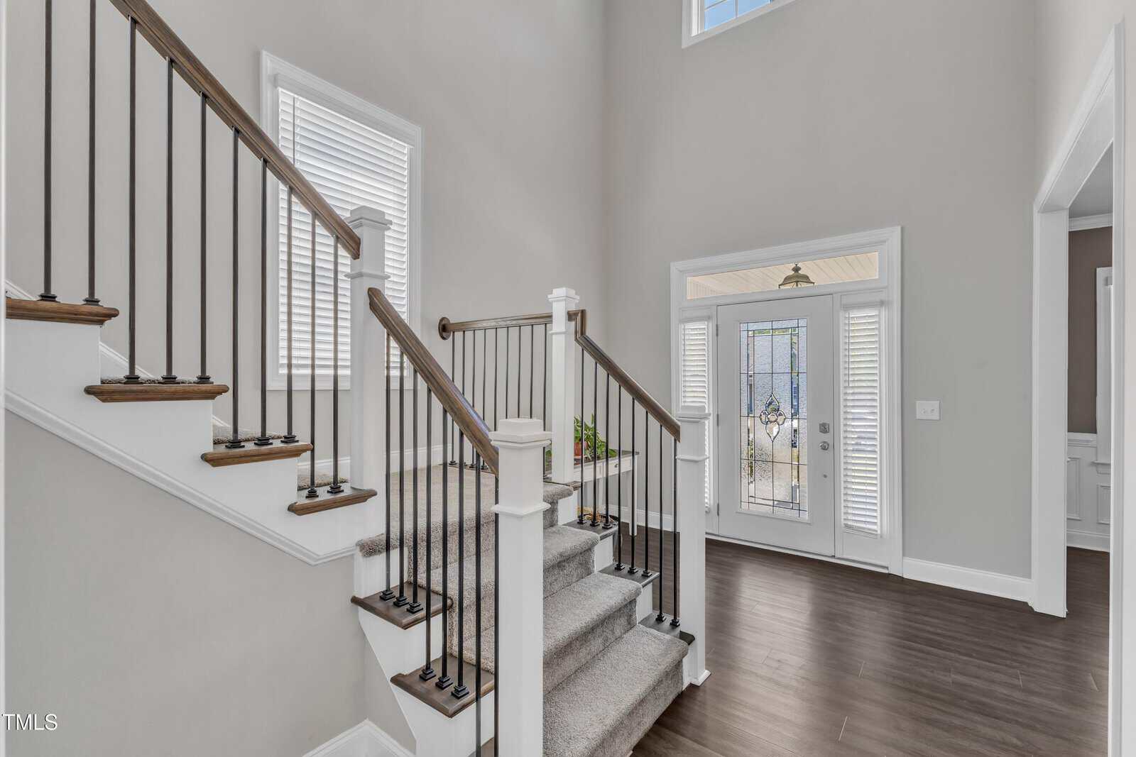 A modern home entryway with a wooden staircase featuring white spindles and carpeted steps, dark wood flooring, light gray walls, and a glass front door with sidelights. Natural light comes in through the windows.