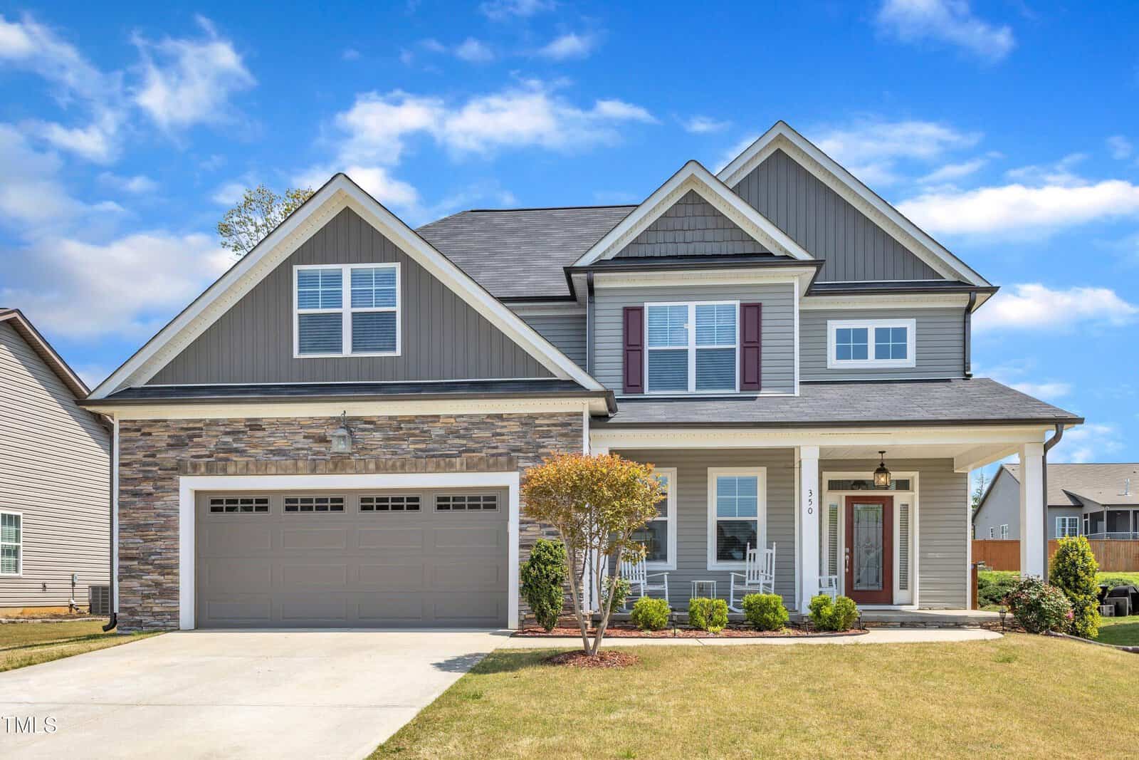 A two-story suburban house with gray siding, stone accents, a double garage, white trim, and a well-kept lawn, photographed under a partly cloudy sky.