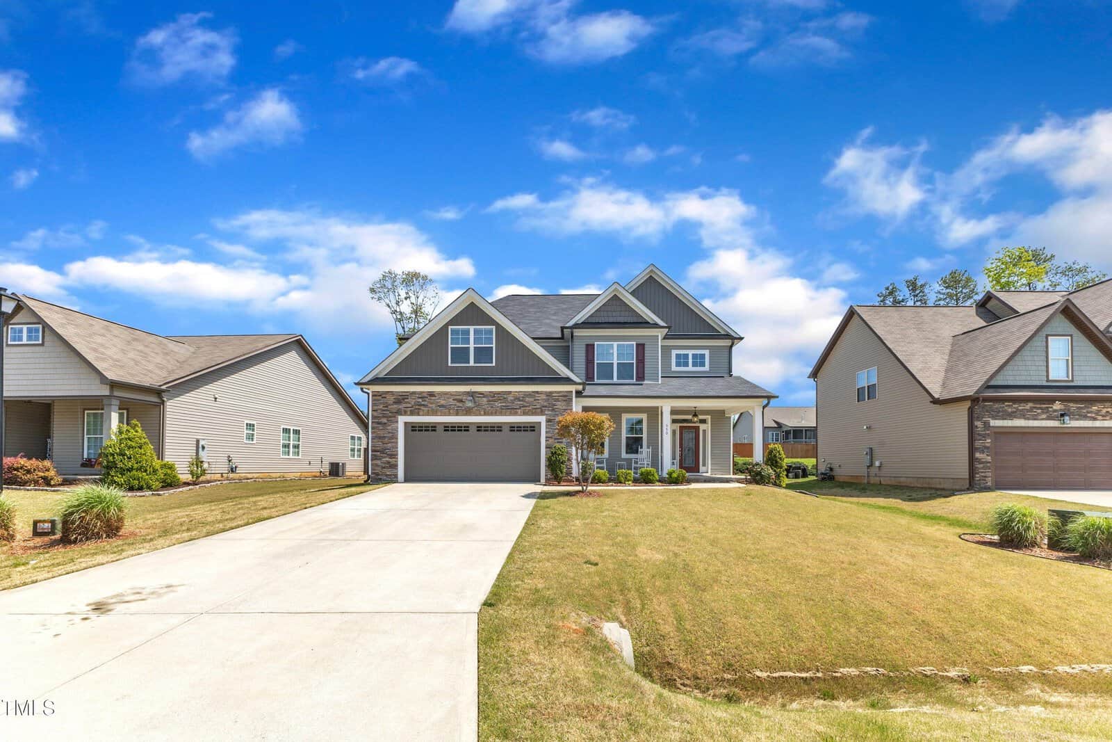 A two-story suburban house with a double garage, gable roof, and a well-maintained front yard sits between two similar neighboring homes under a blue sky with scattered clouds.