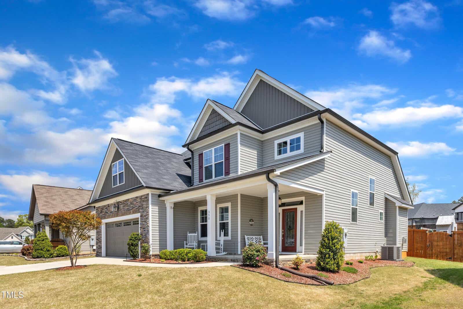 A two-story gray suburban house with white trim, a front porch, attached garage, and manicured landscaping, set under a blue sky with scattered clouds.