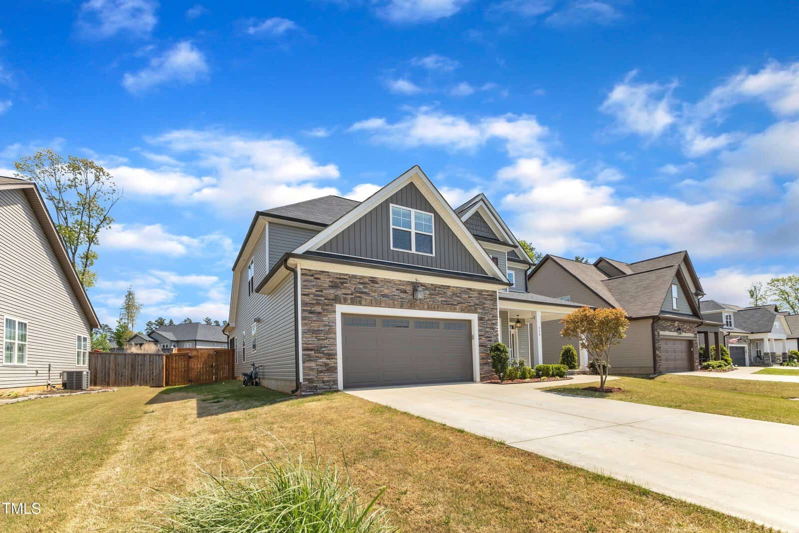 Two-story suburban house with gray siding, stone accents, and a double garage. A concrete driveway leads to the house, and there is a small front porch. The lawn is dry and other houses are visible nearby under a blue sky with clouds.