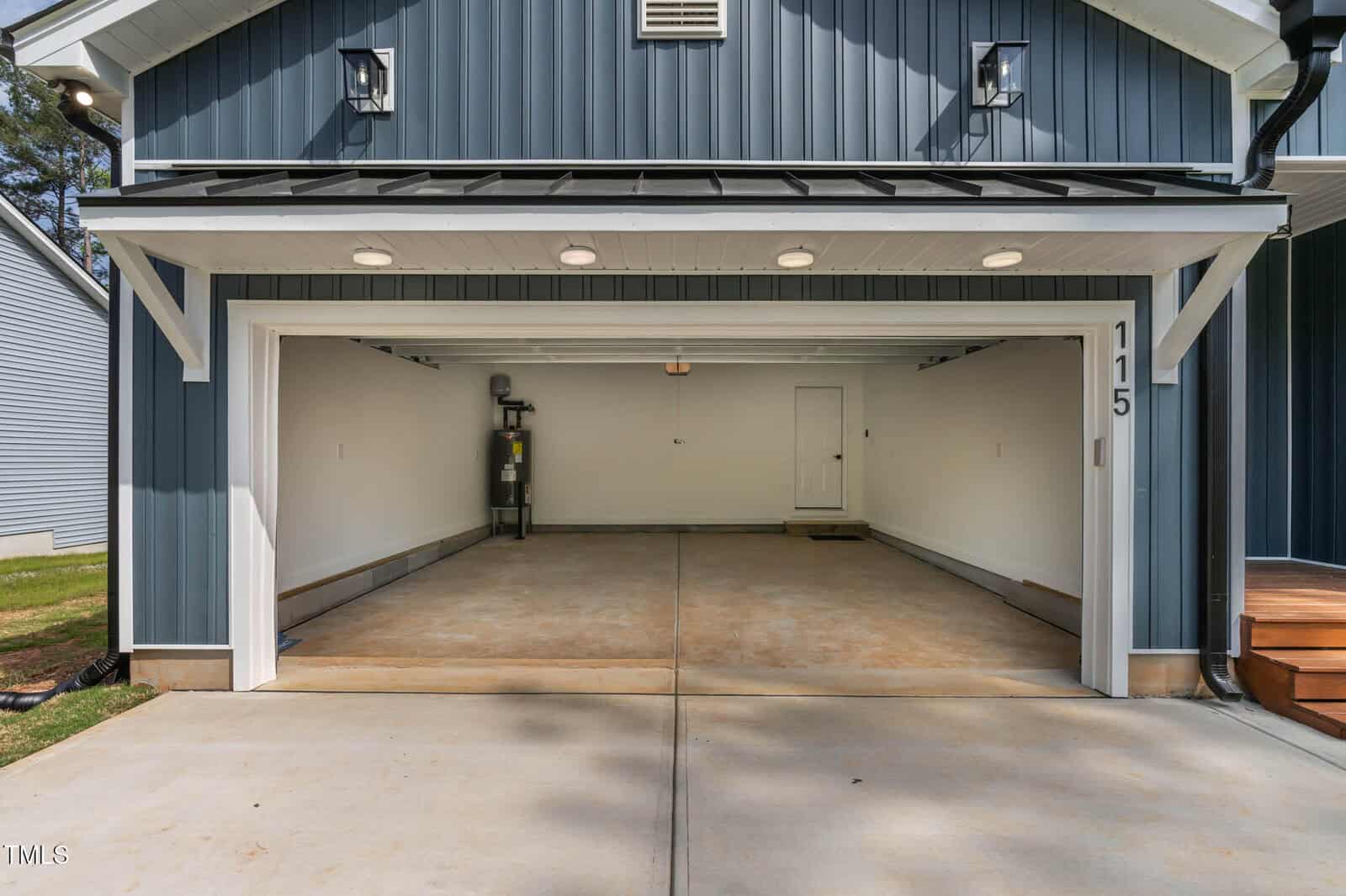 Open two-car garage attached to a blue house, featuring clean concrete floors, white walls, a water heater on the left, and a white door at the back. The address “115” is visible on the right post.