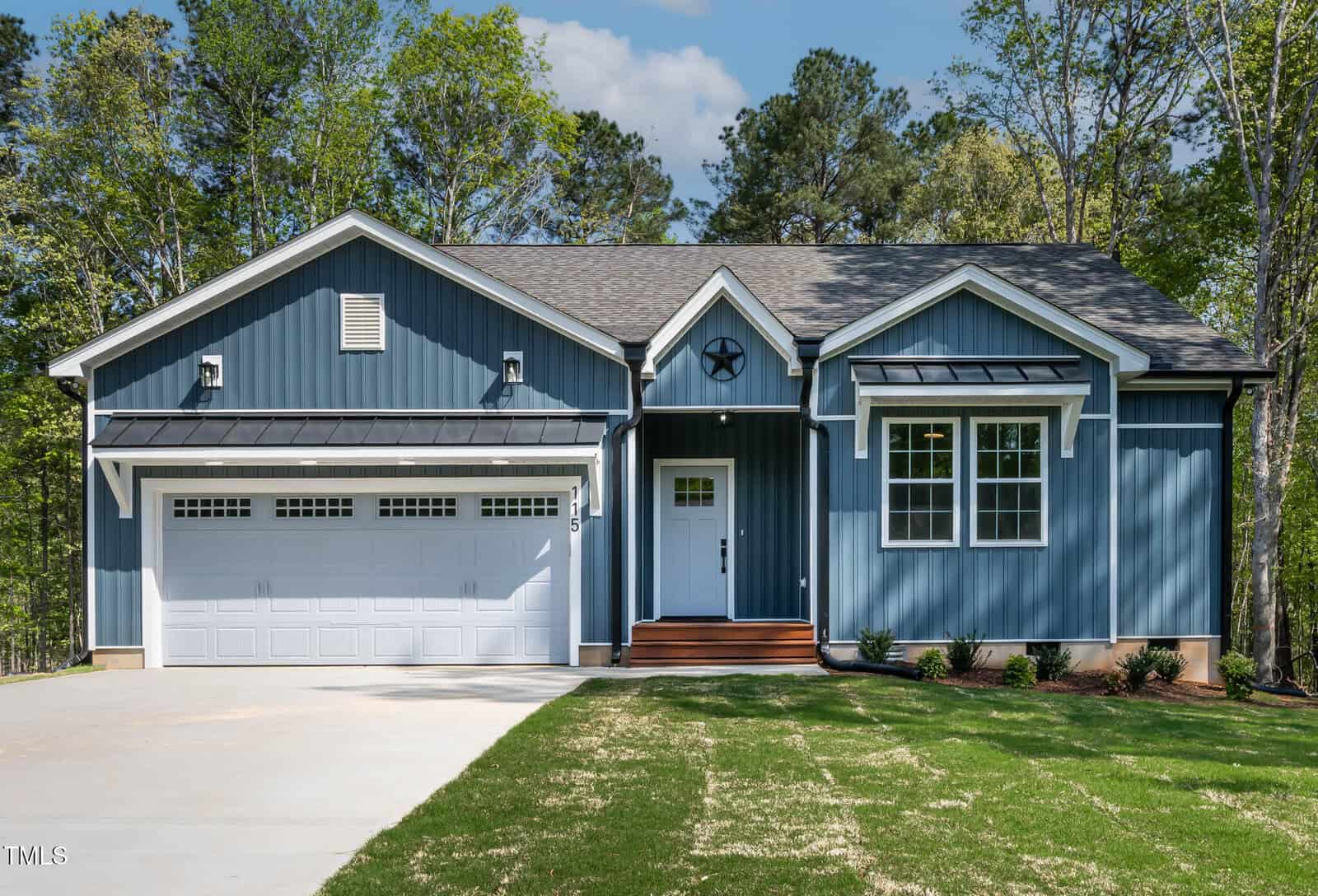 A blue, single-story house with white trim, a two-car garage, front steps, and a manicured lawn, surrounded by trees under a partly cloudy sky sits at 115 Buckskin Drive in Louisburg.