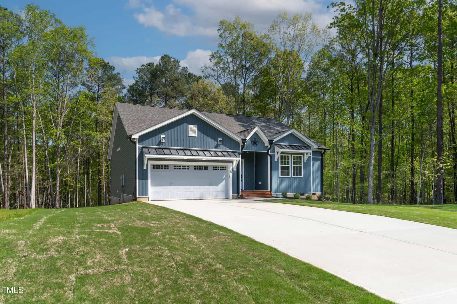 A single-story blue house with a gray roof, white trim, and a two-car garage sits at the end of a wide concrete driveway, surrounded by trees and a partially grassed lawn under a partly cloudy sky.